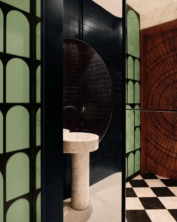 This powder room, attached to an entertainment den in Bangalore, has a bold, moody atmosphere. It features black tiles, checkered flooring, a carved wood door, and a sculptural pedestal marble sink.