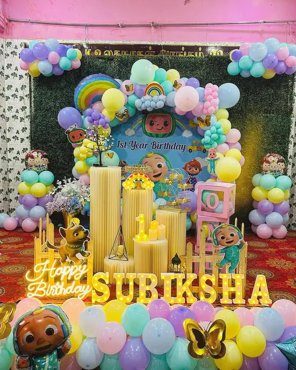 A complete Cocomelon themed first birthday party setup. This package includes a large, vibrant backdrop with characters, an organic balloon garland in pastel colors, Cocomelon foil balloons, and marquee letters spelling out the birthday child's name.
