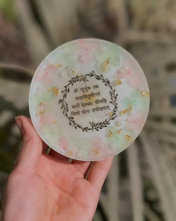 This is one of my favorite pieces, a resin coaster engraved with the Gayatri Mantra. It's surrounded by a delicate floral wreath design and set against a background of soft pastel colors and gold leaf.