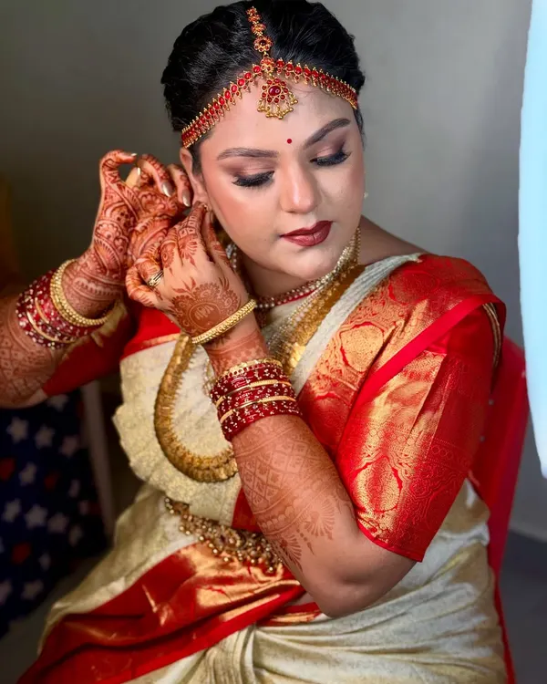 A candid moment of a bride getting ready, putting on her earrings. The makeup is set for a classic South Indian muhurtham look, with defined eyes and a bold lip to match her red and cream silk saree.