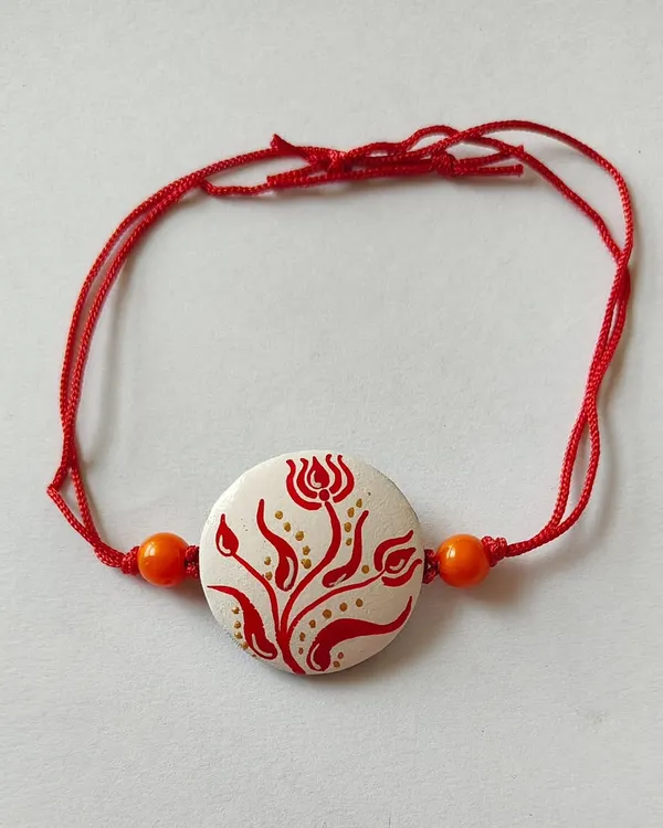 This handmade terracotta rakhi features a delicate red floral design on a white background, inspired by Bengali alpona. It's an elegant and artistic choice for this special festival.