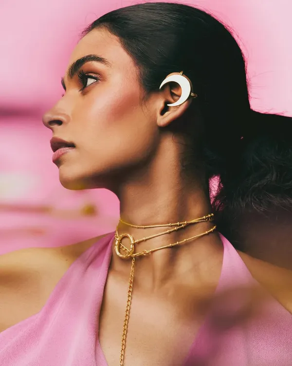 A profile shot for the Batoki Art jewelry campaign, featuring a delicate moon-shaped ear cuff and layered gold necklace. The soft pink background and gentle lighting create a romantic, feminine mood.