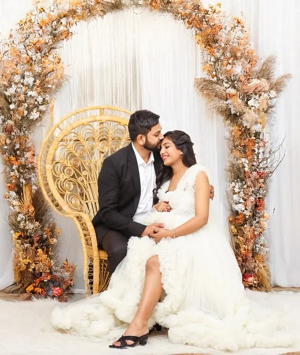 An expecting couple seated in a grand peacock chair, surrounded by a beautiful arch of pampas grass and autumn florals. This dreamy studio setup creates a warm and romantic atmosphere.