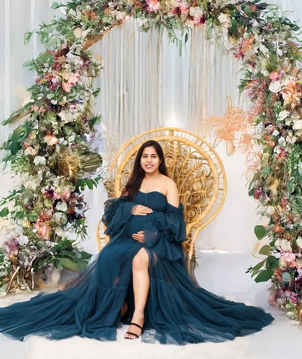 A stunning solo portrait of a mother-to-be seated on a wicker peacock chair, framed by a beautiful floral arch. The rich blue of her gown and the lush flowers create an enchanting and elegant scene.