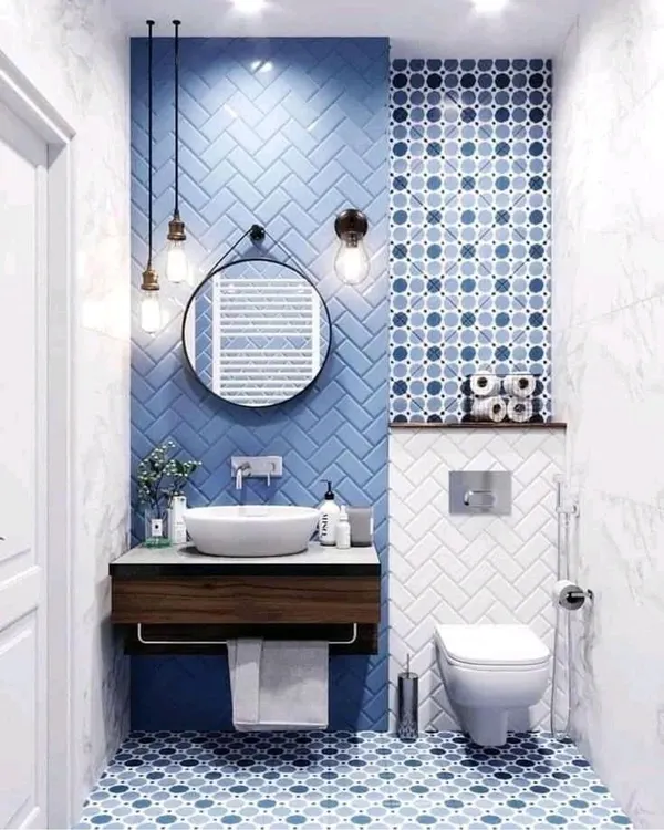 A vibrant and playful bathroom design featuring blue subway tiles in a herringbone pattern and complementary patterned floor tiles. The round mirror and hanging Edison bulbs add a touch of industrial-chic style.