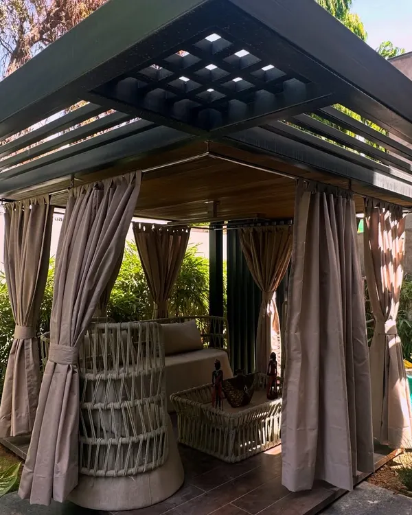 A full view of the custom pergola I designed in Jaipur. It features a slatted roof for partial shade and can be enclosed with curtains, creating a versatile outdoor room surrounded by lush greenery.