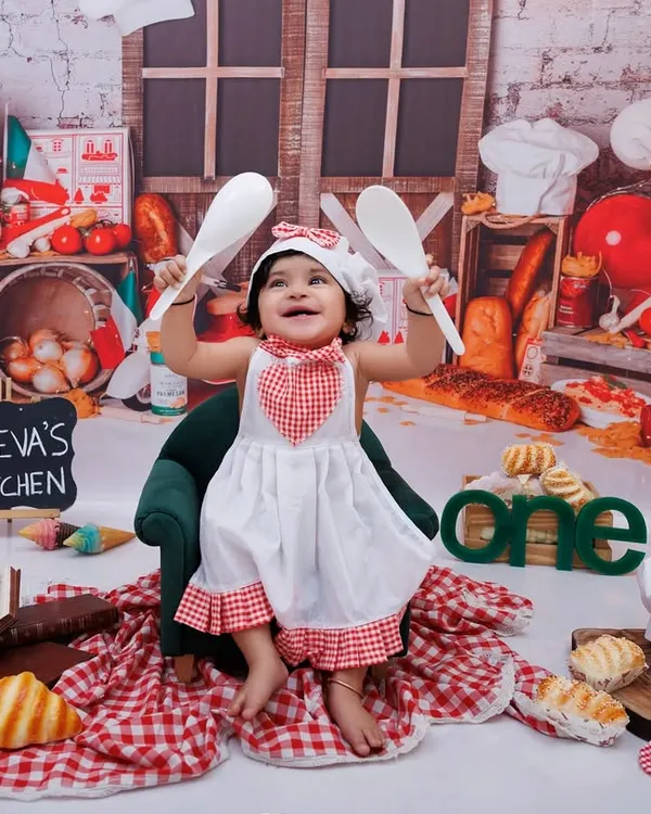 Welcome to Jeeva's Kitchen! This little chef is ready to cook up some fun for her first birthday, holding up her spoons with excitement.