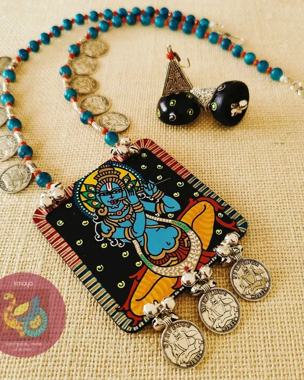 A hand-painted Sri Krishna Murari necklace, showing the lord in a classic pose. The MDF pendant is lacquer polished for a lasting finish and paired with wooden jhumkas, acrylic beads, and metal coin embellishments for a traditional feel.