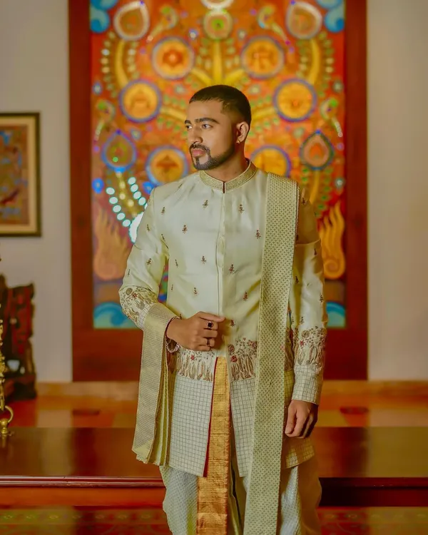This is a custom-designed Indo-Western sherwani set. The off-white jacket features subtle embroidery on the sleeves and body, paired with a traditional dhoti and sash for a look that is both regal and contemporary.