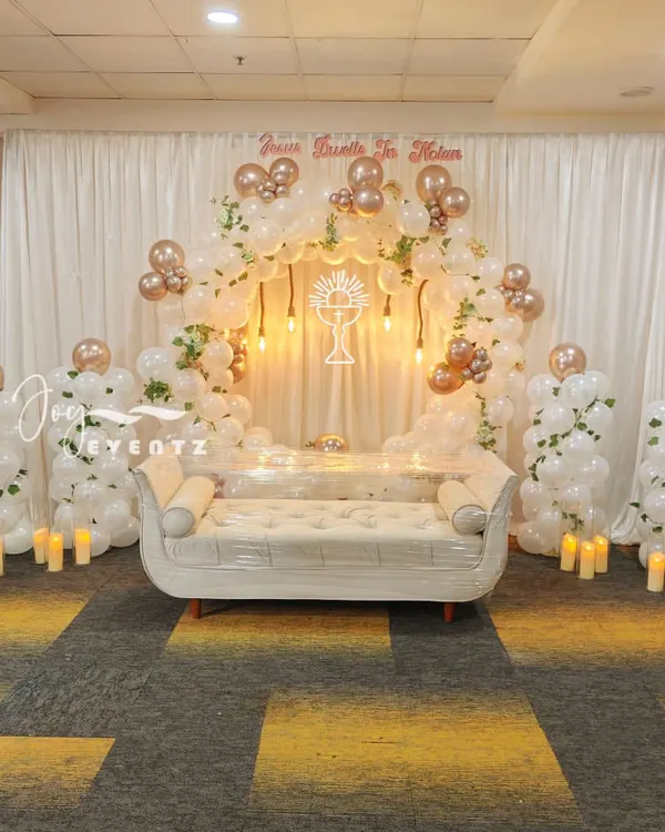 An elegant and expansive stage decoration for a First Holy Communion. This setup features a large balloon arch in white and rose gold, a comfortable white sofa, and a glowing chalice symbol at the center.