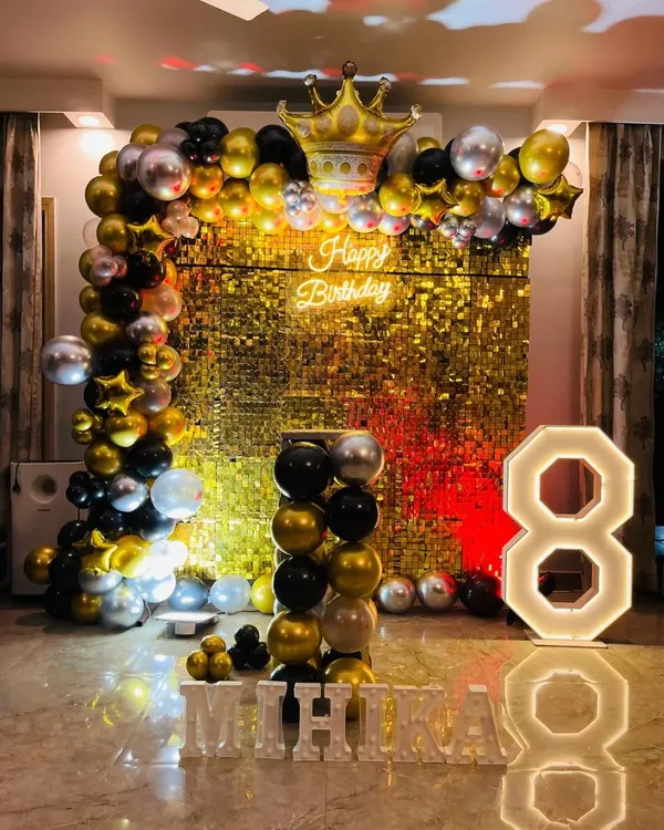 A glamorous 8th birthday with a gold shimmer wall. This stunning setup includes a black, gold, and silver balloon arch, a crown foil balloon, a neon sign, and large light-up letters for the name 'MIHIKA'.