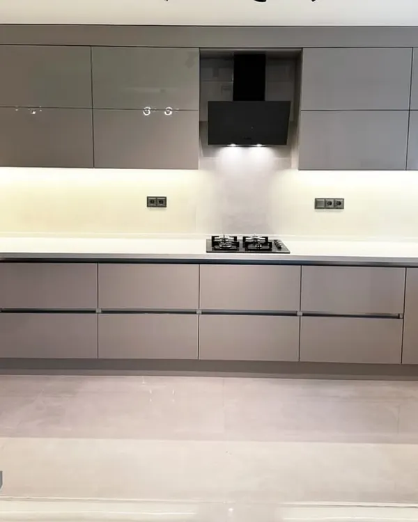 A perfect example of minimalist design in this Gurgaon kitchen, featuring handle-less cabinetry in a glossy beige finish. The continuous under-cabinet LED lighting illuminates the white quartz countertop, creating a clean and spacious feel.
