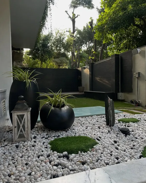 A tranquil Zen garden at the entrance of a Noida home. This design uses a bed of white pebbles, moss patches, and sculptural black planters to create a sense of calm and mindfulness from the moment you arrive.