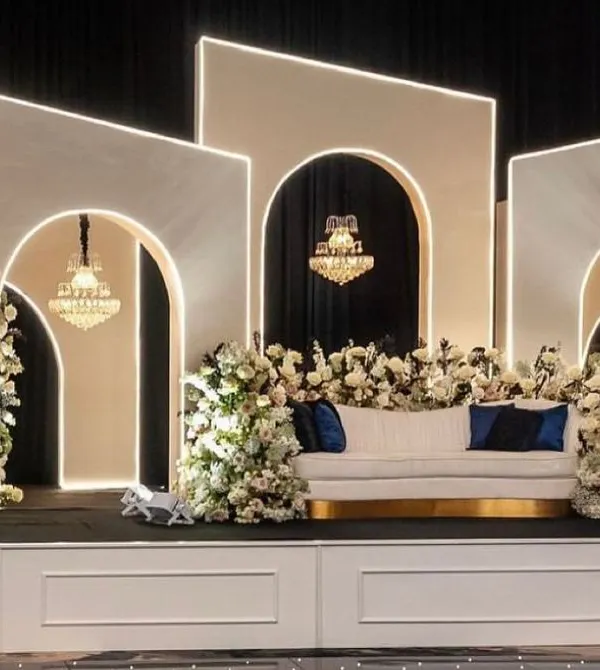 A modern and sophisticated stage design featuring geometric white panels with integrated lighting, grand chandeliers, and lush white floral arrangements for a luxurious feel.
