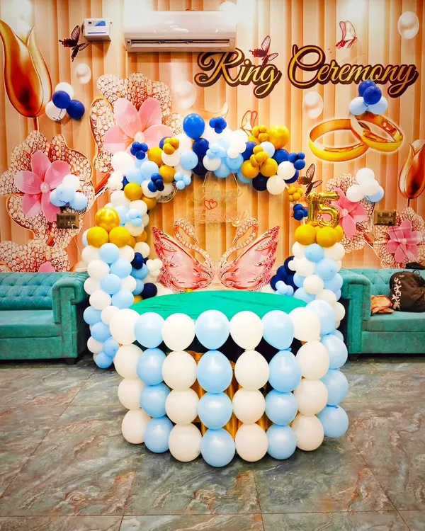 A beautiful and elaborate setup for a ring ceremony. This decoration features a table made of balloons and a colorful balloon arch in blue, white, and yellow, with swan and ring details.