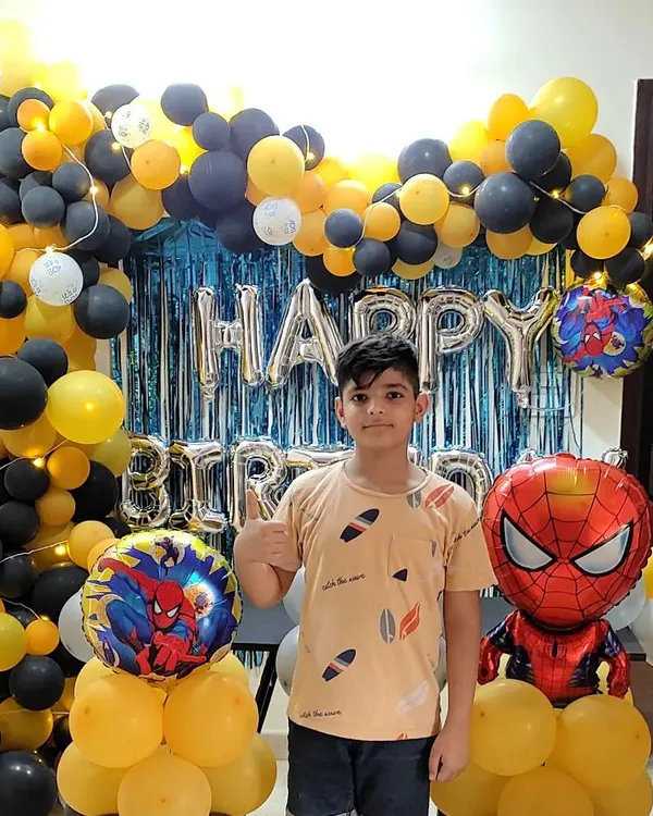 A happy birthday boy posing with his Spider-Man themed party decoration. This setup features a black and yellow balloon arch, a blue foil curtain, and character balloons of the friendly neighborhood superhero.