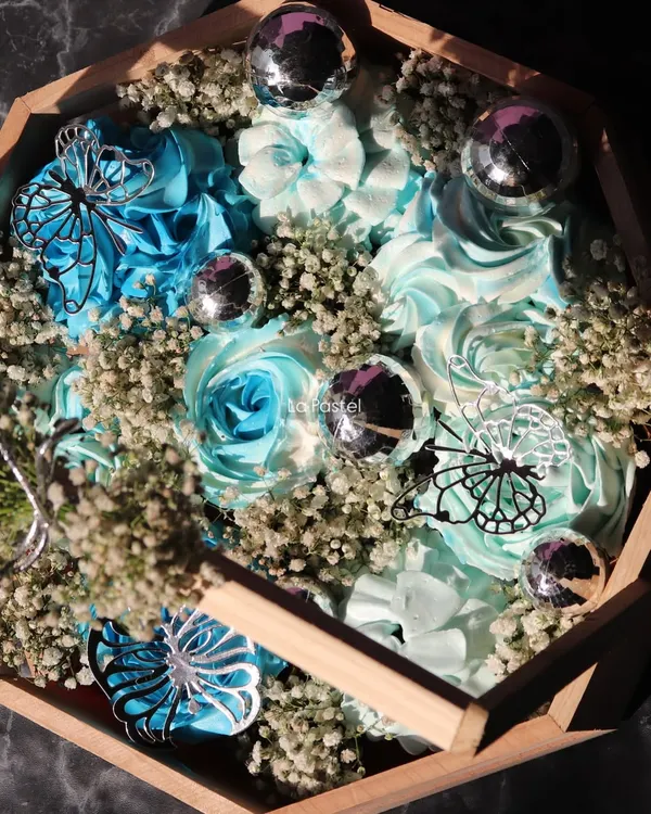 A close-up of the blue-themed cupcake basket, showing the detailed frosting work, silver butterflies, and disco ball accents.