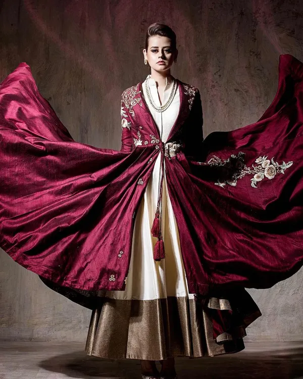 A full-length shot capturing the dramatic flair of a rich maroon embroidered jacket. The movement in the fabric is the central focus, creating a high-impact image for designer campaigns.