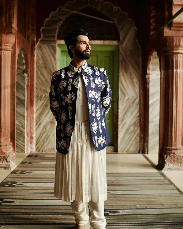 A full-length portrait showcasing a white pleated kurta layered with a navy blue Nehru jacket. The jacket is covered in intricate white floral embroidery, creating a look inspired by period dramas and royal aesthetics.