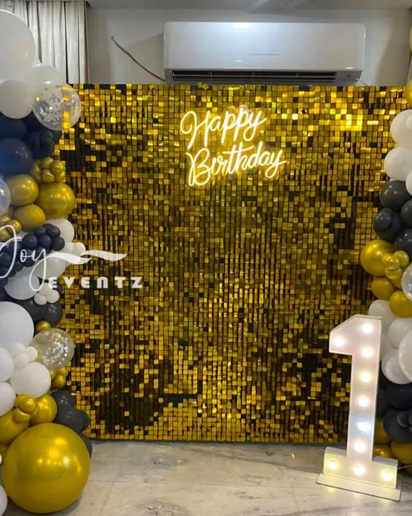 This is a classic and elegant first birthday setup featuring a gold shimmer wall. The backdrop is complemented by a 'Happy Birthday' neon sign and a beautiful balloon garland in black, white, and gold.
