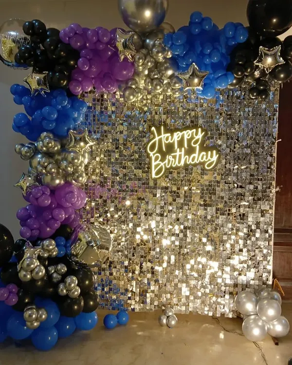 A vibrant party setup with a silver sequin wall. The backdrop is framed by a balloon garland in blue, purple, and black, and features a 'Happy Birthday' neon sign for a disco-ready vibe.