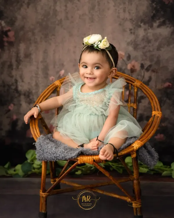 A perfect sitter session portrait. This happy baby girl is smiling brightly while sitting in a small wicker chair against a classic, painterly backdrop.