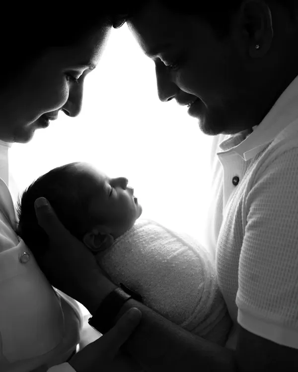 An intimate black and white silhouette of both parents gazing down at their newborn baby held lovingly in their hands.