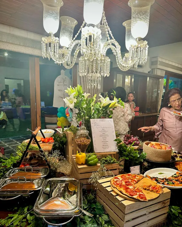 An elegant indoor setup for a house party. The buffet table is lavishly decorated with a crystal chandelier overhead, fresh white lilies, and our signature wooden crates, creating a luxurious feel while guests enjoy fresh pizza and other dishes.