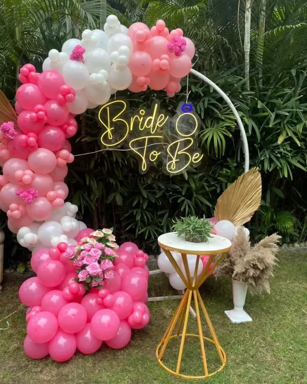 We created this chic 'Bride to Be' backdrop for an outdoor celebration. It features a custom neon sign on a white circular frame, wrapped in a vibrant pink and white balloon garland with decorative palm leaves.
