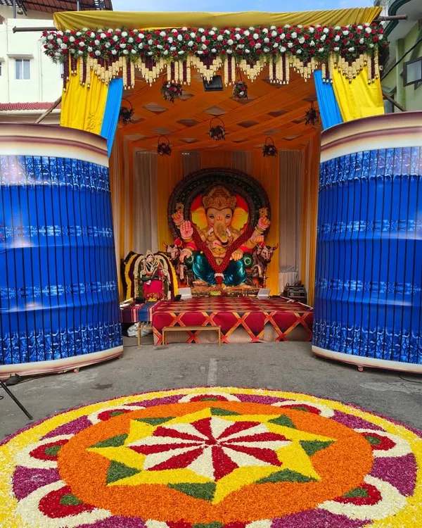 This is a large-scale outdoor pandal we constructed for a Ganesh Chaturthi festival celebration. The setup includes a decorated stage for the idol, vibrant yellow and blue fabric draping, and a massive, colorful flower rangoli on the ground.