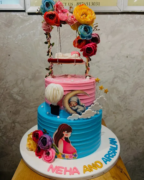 A beautiful two-tier baby shower cake with pink and blue layers, featuring a sleeping baby on a swing, a pregnant mother illustration, and floral details.