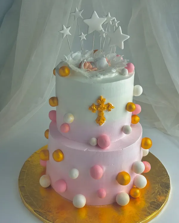 A beautiful two-tier cake for a child's dedication ceremony. The design features a sleeping baby in an angel's nest, a golden cross, and soft pink and white spheres, symbolizing a precious gift from above.