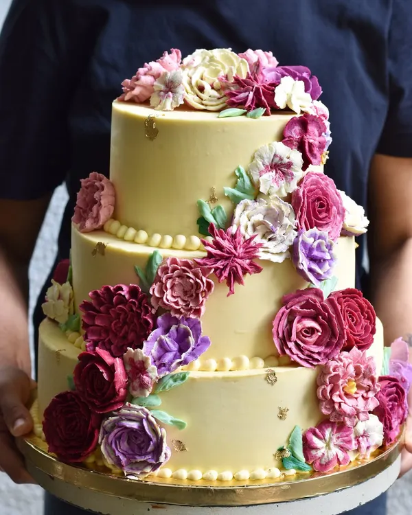 A stunning three-tiered wedding cake featuring a cascade of hand-piped buttercream flowers in shades of purple and pink. We specialize in creating these showstoppers that taste even better than they look.