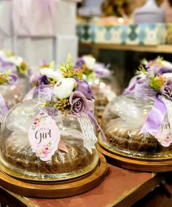 A close-up of our popular baby girl announcement favors. Each cake is presented under a protective dome and beautifully decorated with a posy of purple and white flowers, a lace ribbon, and a custom "It's a Girl" tag, making for a truly special keepsake gift.