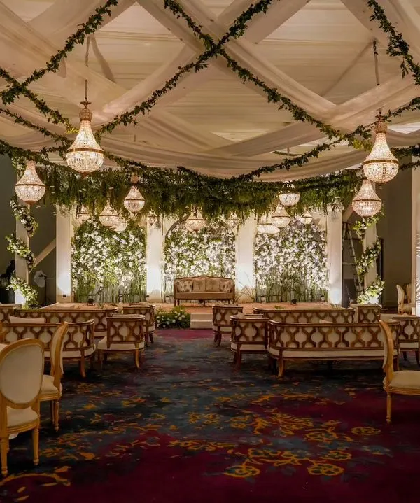 A breathtaking reception hall designed as an enchanted forest. The ceiling is draped with fabric and hanging greenery, with crystal chandeliers providing a warm, magical glow over the elegant seating arrangement.