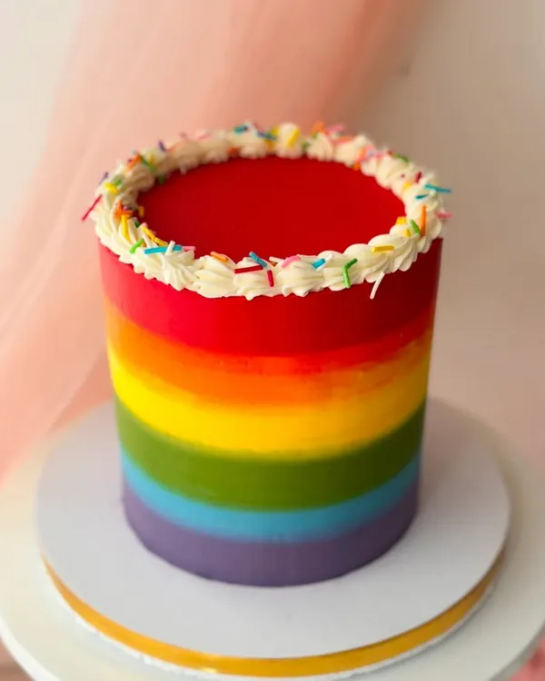 A vibrant rainbow cake with a bright red top. The smooth gradient is created with buttercream, and the top is finished with a simple piped border and sprinkles.