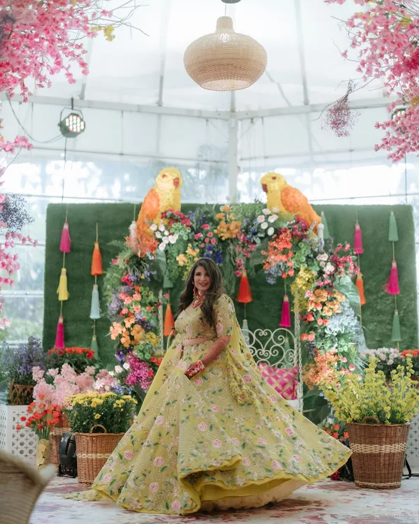 A perfect Mehndi look. The bride is surrounded by a riot of color, and her makeup is fresh and vibrant, with a focus on glowing skin and a bright lip to match her floral lehenga.