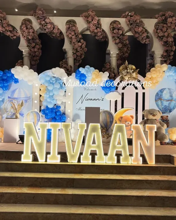 A grand stage setup for Nivaan's welcome party and Mundan ceremony. The design features a hot air balloon theme with large marquee letters, teddy bear props, and an elaborate backdrop with blue and white balloons.