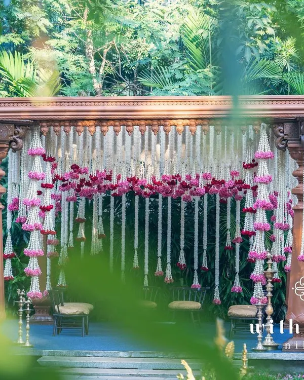 A breathtaking view of our summer wedding mandap, framed by lush greenery. The structure, made of intricately carved South Indian pillars, is adorned with cascading garlands of tuberose and pink oleander, creating a truly romantic and sacred space.