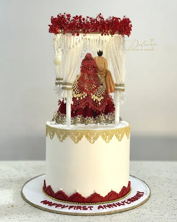 Our full Mandap cake, designed for a first wedding anniversary. The intricate topper, featuring a bride and groom under a floral canopy, sits atop a classic white cake with gold and red lace details.