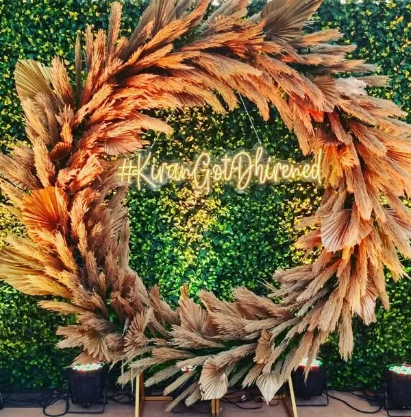 A rustic and chic photo booth for an engagement or reception. The backdrop features a large wreath made of pampas grass against a green wall, with a custom neon hashtag for the couple.