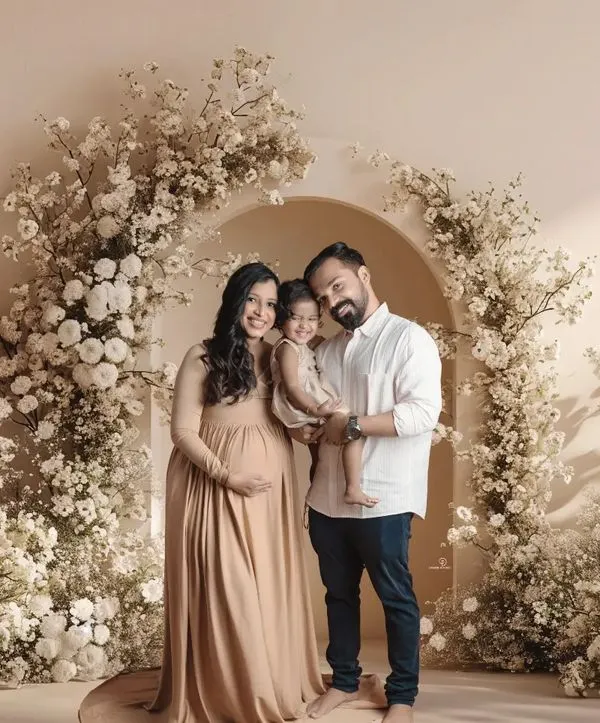 A beautiful family portrait during a maternity session. The expecting parents and their toddler stand together under a stunning arch of white flowers, celebrating the upcoming arrival.