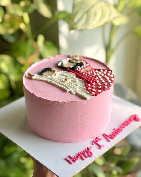 A side view of the first anniversary cake, showing the hand-piped illustration of the couple in their wedding attire on a smooth pink buttercream canvas.