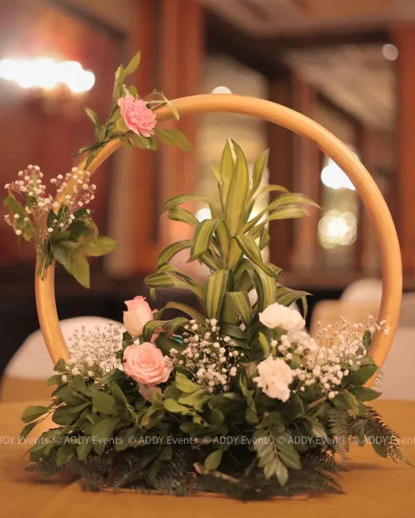 This modern centerpiece features a golden circular frame adorned with soft pink roses, white carnations, and baby's breath. It is a perfect example of how we blend contemporary design with classic floral beauty for table decor.