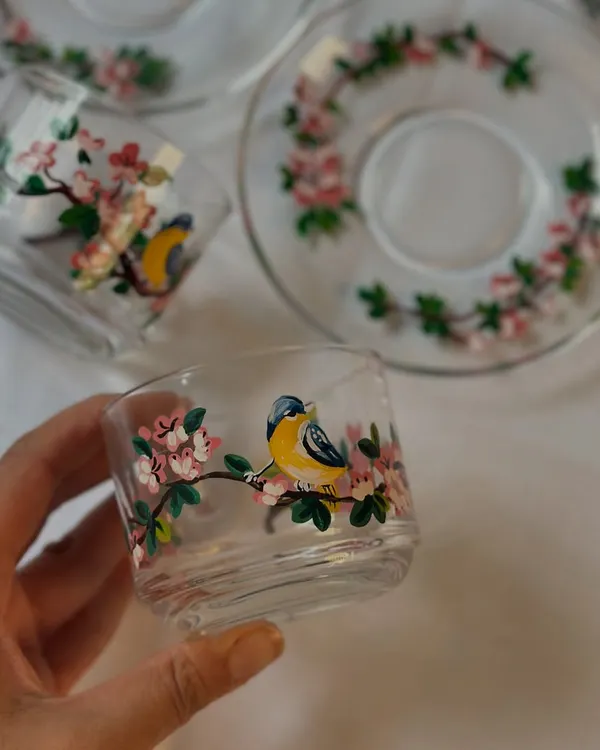 A close-up of our hand-painted glass teacup featuring a delicate yellow bird perched on a cherry blossom branch. Each detail is painted with fine brushes to bring the scene to life.
