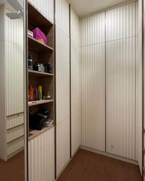 A walk-in closet space featuring floor-to-ceiling wardrobes with a unique curved fluting design on the doors. The design incorporates both closed storage and open shelving for display.