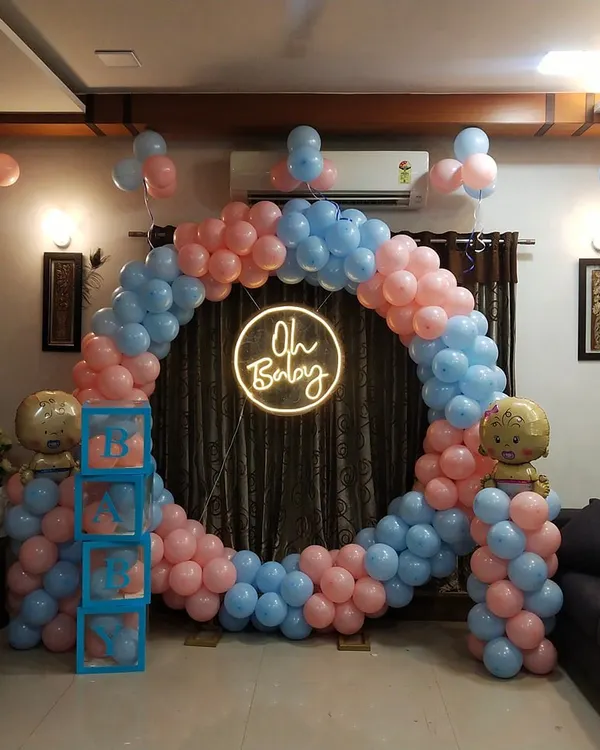 A wider view of the at-home baby shower setup, showing how the large circular balloon arch fits perfectly within a living room. The design includes 'BABY' blocks and balloon pillars with baby-themed foils.