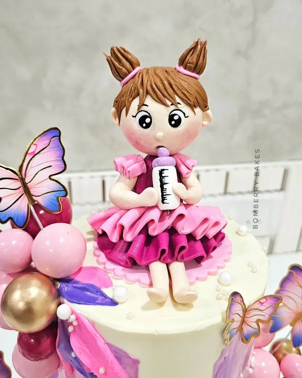 A close-up of a handmade fondant figurine for a first birthday cake. I teach how to sculpt expressive faces, create detailed clothing like this ruffled pink dress, and add small accessories like the milk bottle.