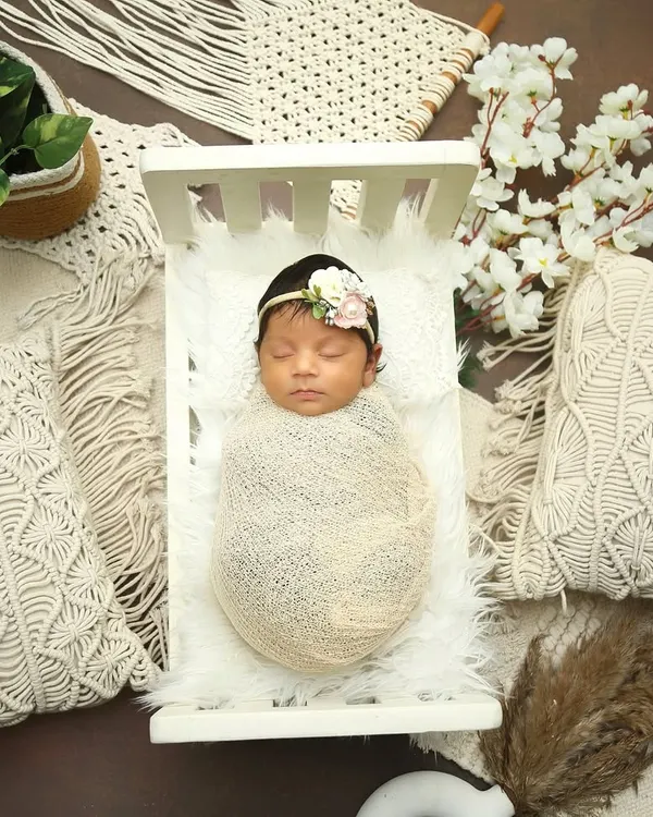 A beautifully styled shot featuring a newborn in a miniature white crib. The boho theme with macrame pillows and pampas grass creates a soft, neutral, and trendy look.
