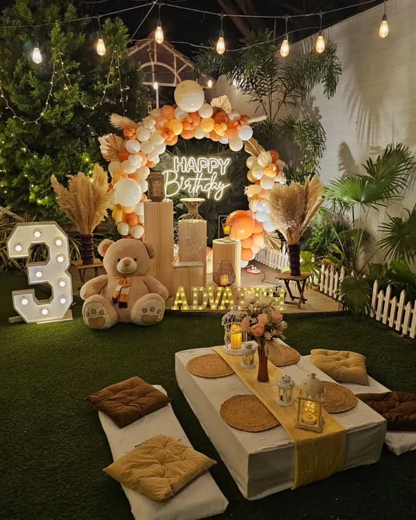 A cozy and chic teddy bear picnic theme for a 3rd birthday. The outdoor setup includes low seating with cushions, a neutral-toned balloon arch, and a light-up number.